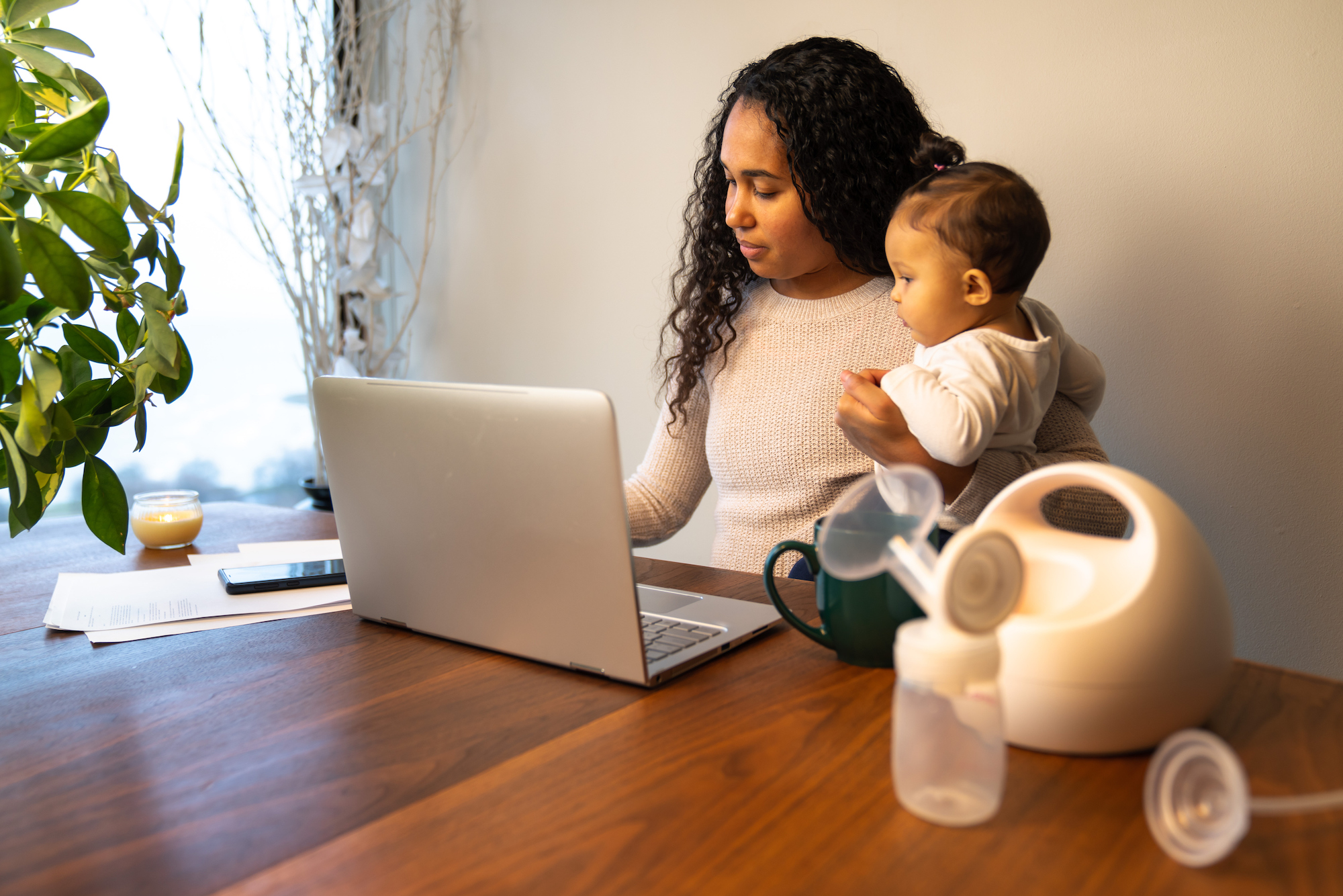 A beautiful young mixed race African American mother holds her daughter while taking notes at her dining table serving as a temporary remote work from home station with breast pump in foreground.