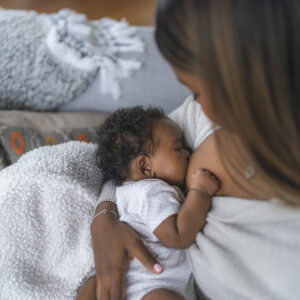 A woman of Indian descent is breastfeeding her baby daughter. The woman is sitting on a couch in her home's living room. The relaxed mother is enjoying the skin to skin contact and bonding time with her child. The image is shot over the woman's shoulder looking down at the baby and is focused on the baby. Family moments and postpartum concepts.