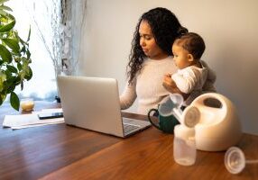 A beautiful young mixed race African American mother holds her daughter while taking notes at her dining table serving as a temporary remote work from home station with breast pump in foreground. A beautiful young mixed race African American mother holds her daughter while taking notes at her dining table serving as a temporary remote work from home station with breast pump in foreground.