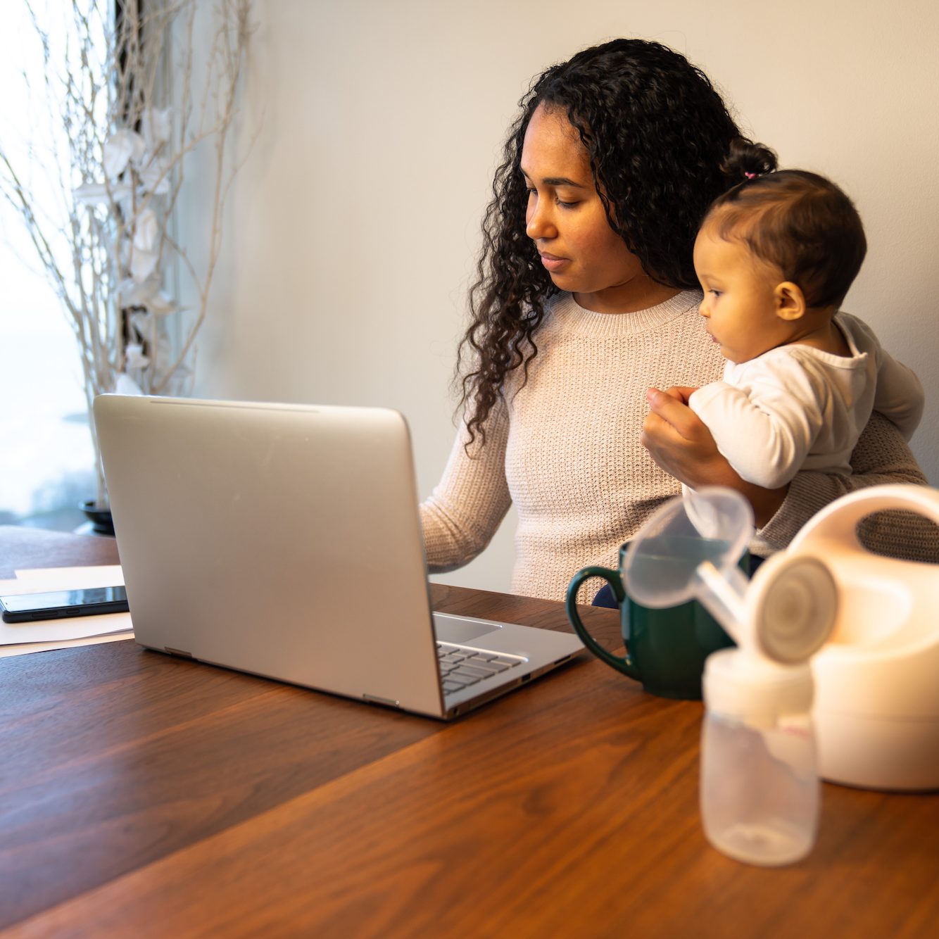 A beautiful young mixed race African American mother holds her daughter while taking notes at her dining table serving as a temporary remote work from home station with breast pump in foreground.